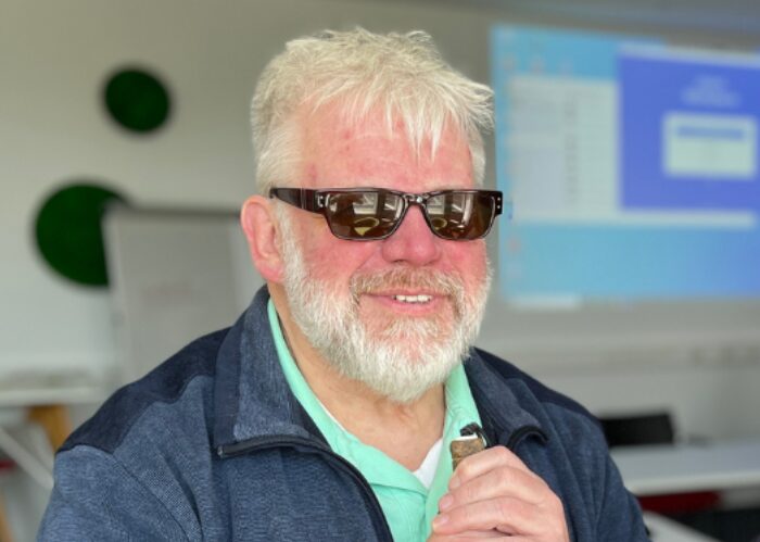 Johannes Willenberg mit Sonnenbrille und einem Gegenstand in der Hand. Im Hintergrund ist ein Büro erkennbar